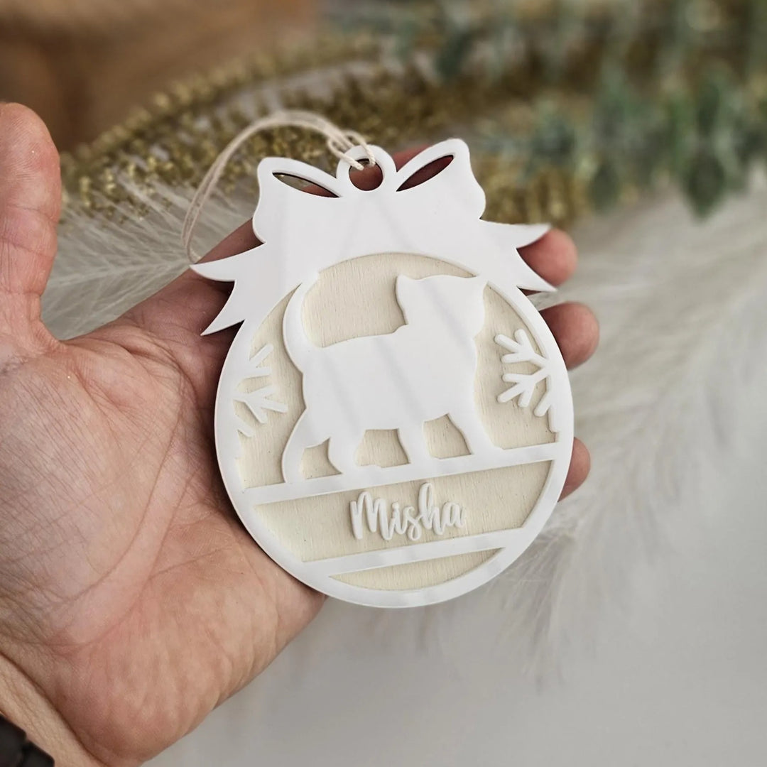 A hand holding a white ornament with a cat silhouette and the name "Mishka" written on it.