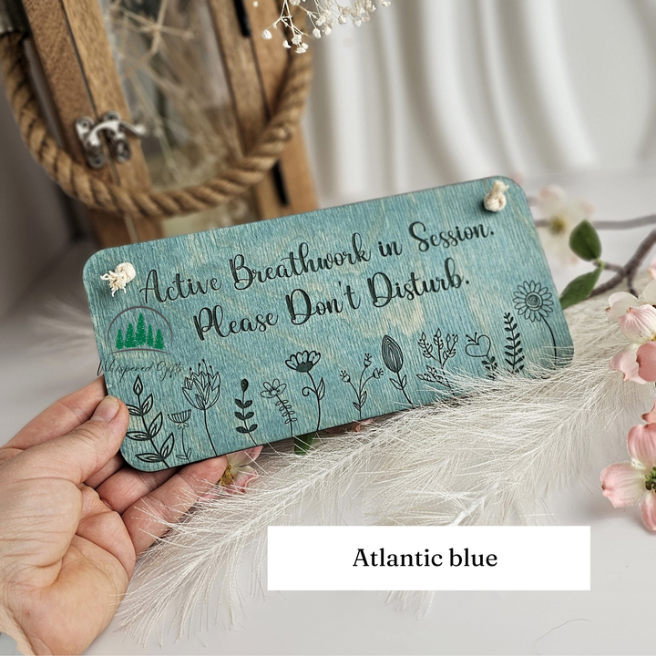 A hand holding a wooden sign with the text "Active Breathwork in Session. Please Don't Disturb." The sign is surrounded by feathers and flowers, and the background includes a wooden lantern and a white curtain.