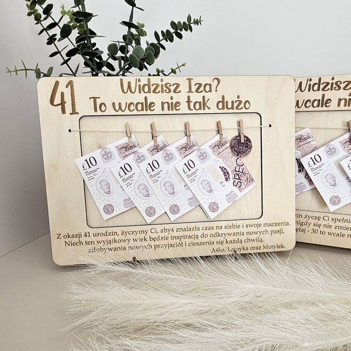 A wooden frame with money notes attached, displaying the text "41 Widzisz Isza? To wale nie tak duzo" and a plant in the background.