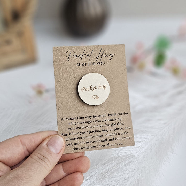 A person's hand holding a small wooden pin with a message about a "Pocket Hug" on a card.