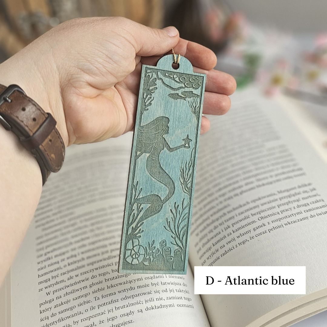 A hand holding a teal-colored mermaid-themed bookmark is resting on an open book.