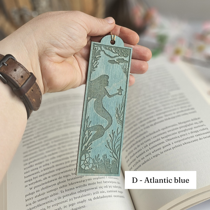 A hand holding a teal-colored mermaid-themed bookmark is resting on an open book.