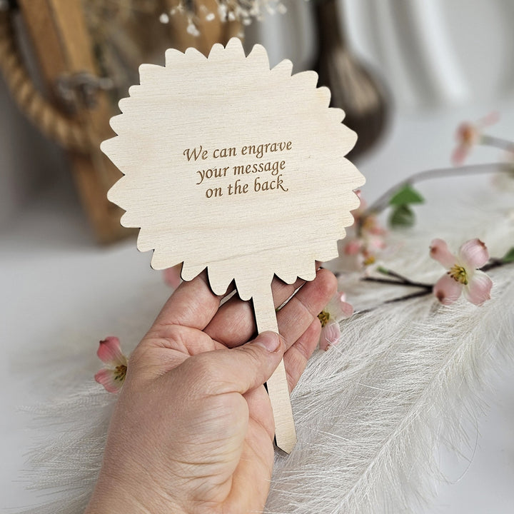 A hand holding a wooden sign with the message "We can engrave your message on the back" against a background of flowers and a wooden frame.