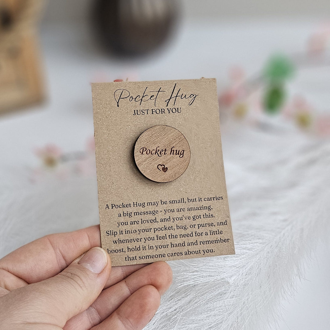 A hand holding a small wooden "Pocket Hug" magnet with a heart-shaped cutout.