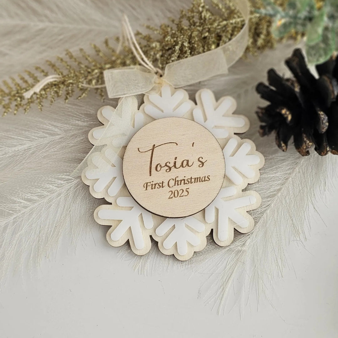 A wooden ornament with a snowflake design, featuring a personalized message that says "Tosia's First Christmas 2025".