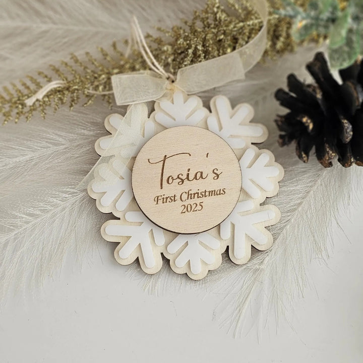 A wooden ornament with a snowflake design, featuring a personalized message that says "Tosia's First Christmas 2025".