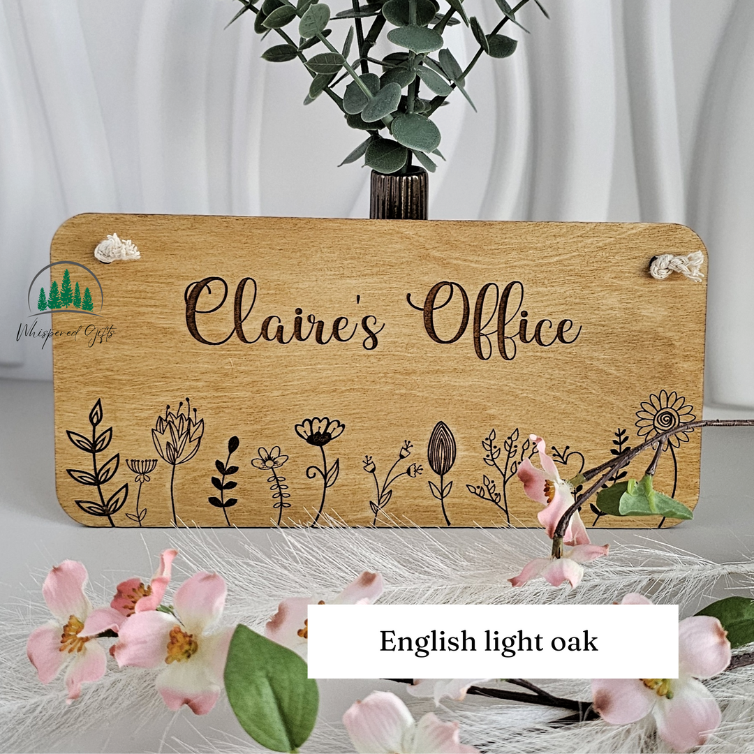 A wooden sign with the name "Claire's Office" written on it, surrounded by various floral designs. The sign is placed on a table with a vase of flowers and greenery in the background.
