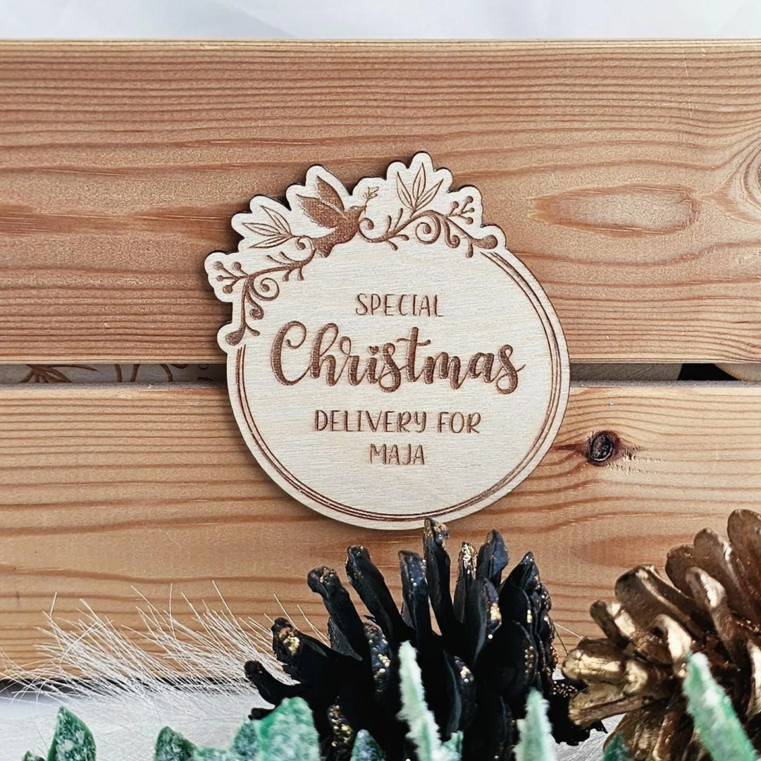 A wooden sign with the text "Special Christmas Delivery for Maja" is displayed on a wooden surface, surrounded by pine cones and other holiday decorations.